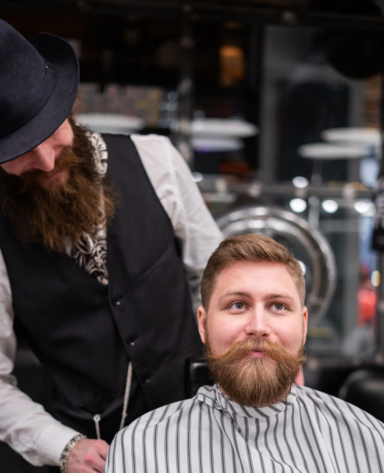 A barber in a hat and waistcoat standing behind a customer in a striped cape